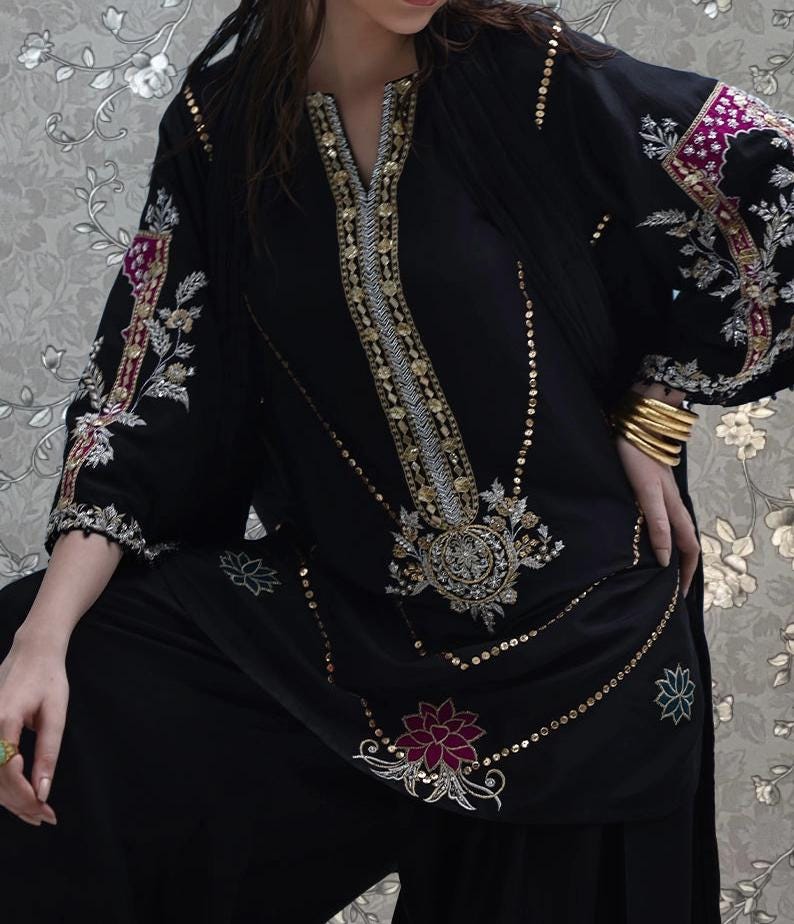A woman wearing a black dress with intricate gold embroidery and floral designs, posing against a floral background.