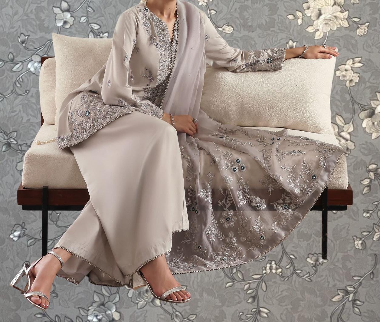 A woman wearing a long, beige dress with intricate embroidery is sitting on a white couch against a gray floral background.