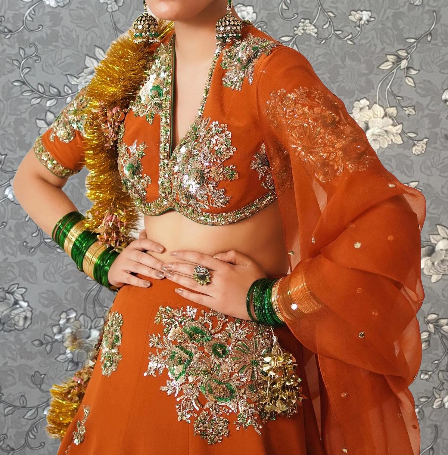 A woman wearing an ornate, colorful dress with intricate embroidery and jewelry poses against a gray floral background.