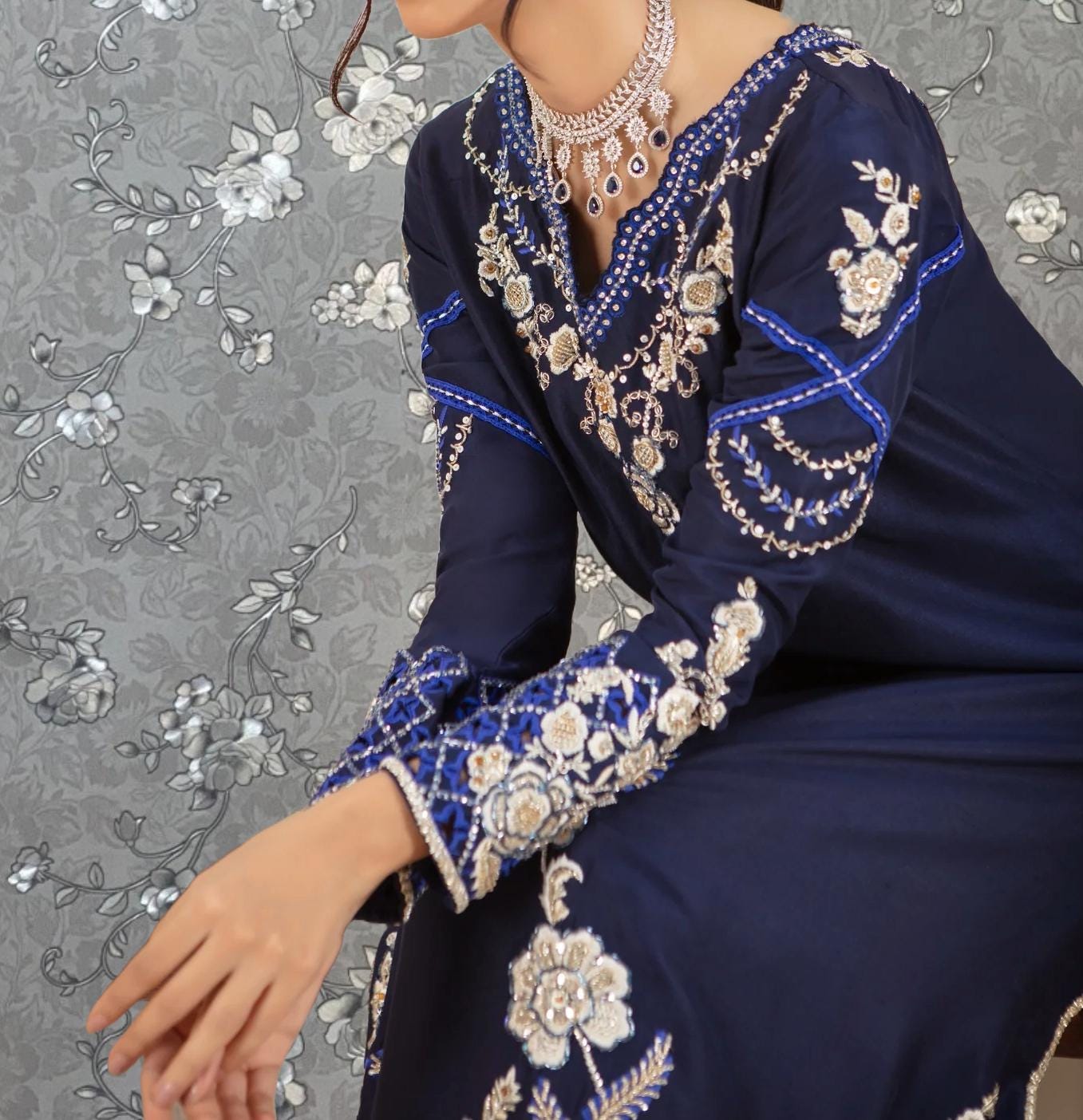 A woman wearing a blue dress with intricate embroidery and jewelry poses against a gray floral background.