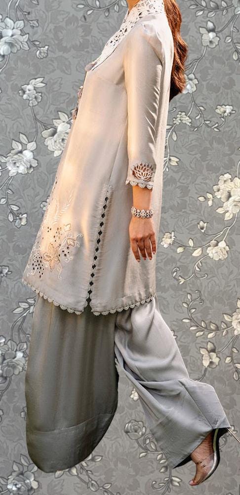 A person wearing a long-sleeved white blouse with intricate embroidery and a matching skirt, standing against a gray floral background.