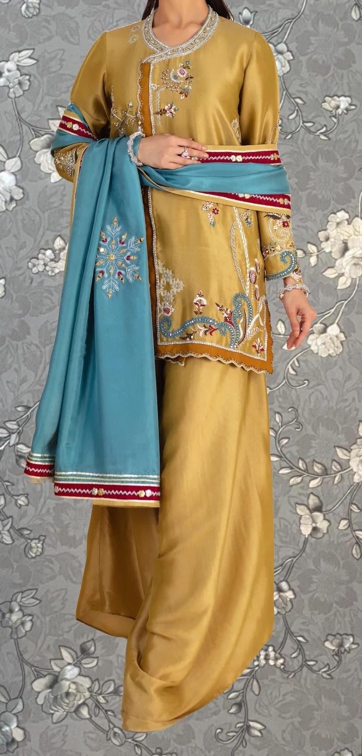 A woman wearing a gold and blue dress with intricate embroidery, standing against a gray floral background.