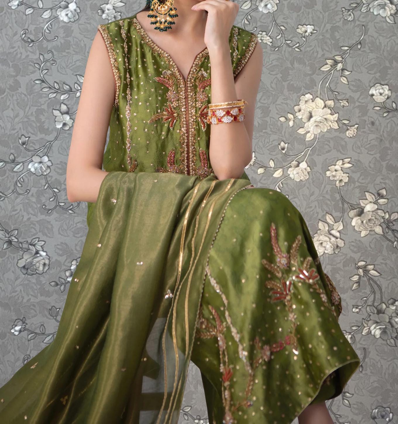 A woman wearing a green and gold dress with intricate embroidery poses against a gray floral background.