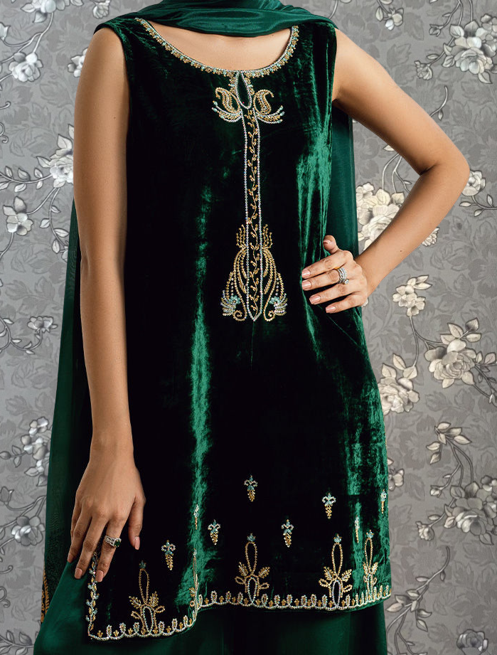A woman wearing a green velvet dress with gold embroidery and floral designs, posing against a gray floral background.