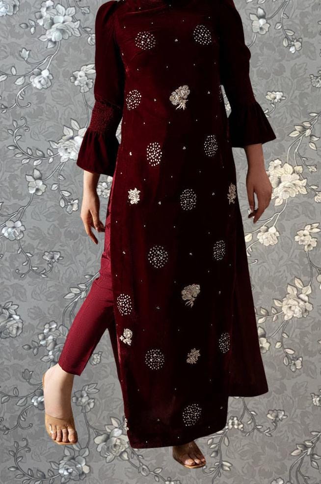 A person wearing a long burgundy dress with white floral embroidery stands against a gray floral background.