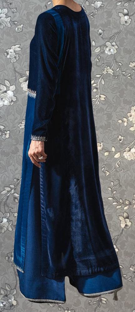 A person wearing a long, blue velvet dress with a high collar and long sleeves stands against a gray floral background.