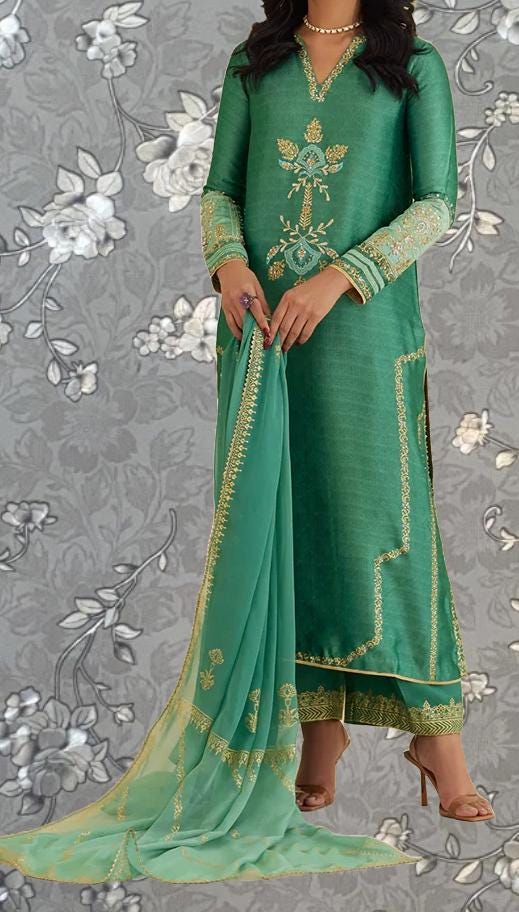 A woman wearing a green and gold dress with intricate embroidery stands against a gray floral background.