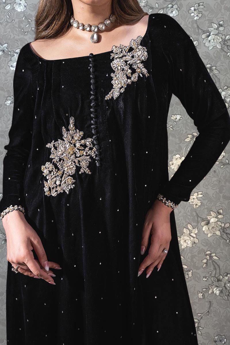 A woman wearing a black dress with intricate embroidered designs and jewelry poses against a floral background.