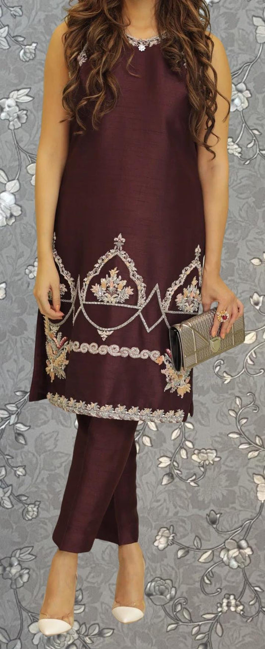 A woman wearing a burgundy dress with intricate embroidery and holding a small clutch purse stands against a gray floral background.