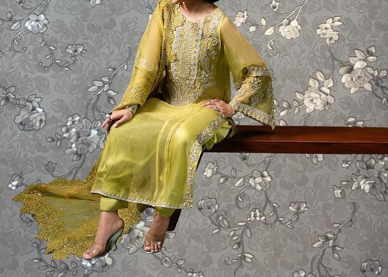 A woman wearing a yellow and gold dress with intricate embroidery is sitting on a wooden bench against a gray floral wallpaper background.