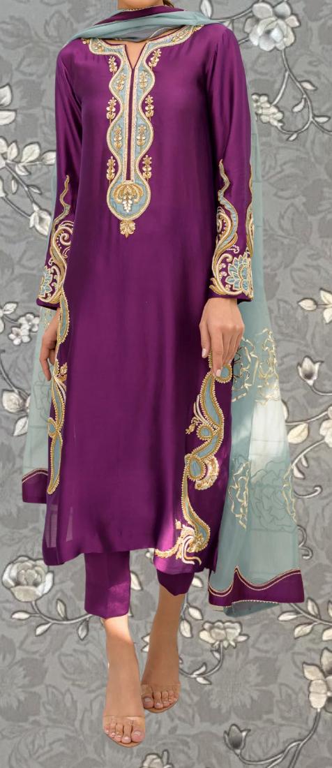 A person wearing a purple and green dress with intricate embroidery and patterns, standing against a gray floral background.
