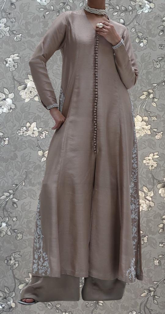 A person wearing a long, beige dress with floral embroidery stands against a floral background.