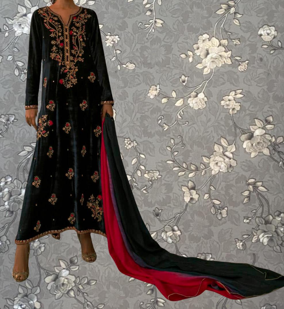 A person wearing a long black dress with gold embroidery and a red and black shawl draped over their shoulders, standing against a gray floral background.