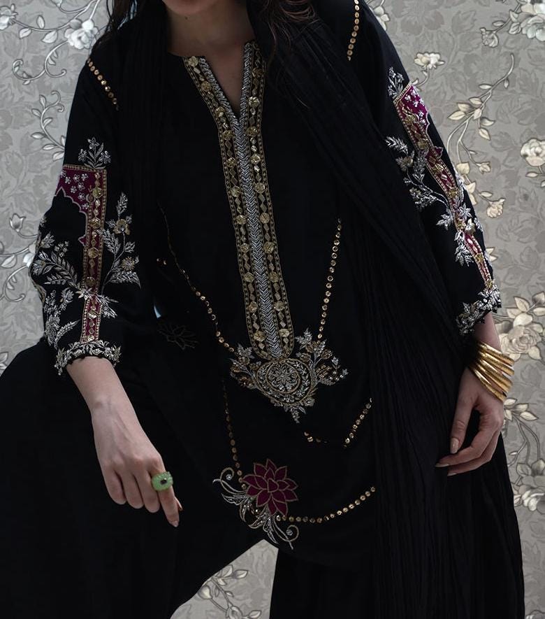 A person wearing a black dress with intricate embroidery and gold accents, posing against a floral background.