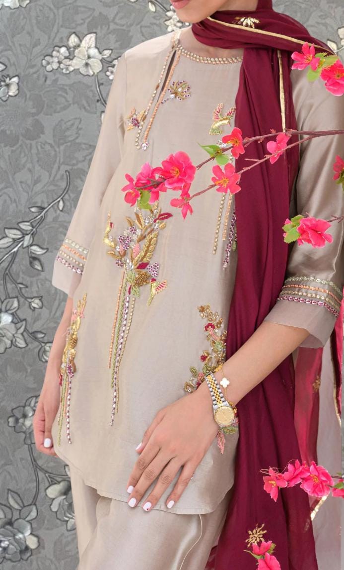 A woman wearing a beige dress with intricate floral embroidery and a burgundy scarf, posing against a gray floral background.