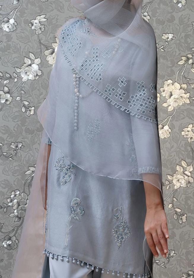 A person wearing a gray dress with intricate embroidery and a sheer overlay, standing against a floral patterned background.