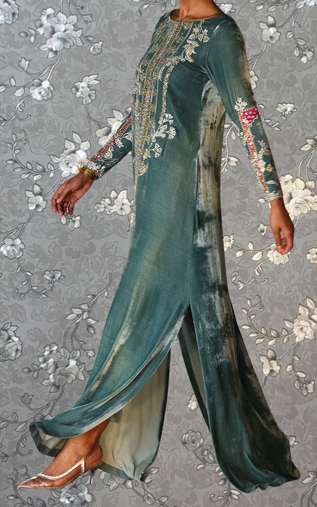 A woman wearing a long, green dress with intricate embroidery and floral patterns, standing against a gray floral background.