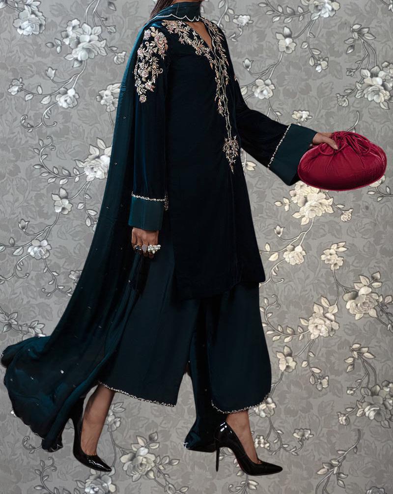 A person wearing a long black dress with gold embroidery and black high heels is holding a red purse against a gray floral background.