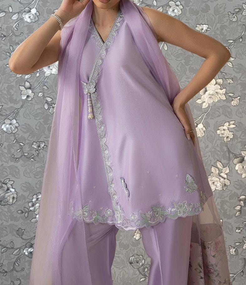 A woman wearing a lavender dress with a floral pattern and a matching scarf stands in front of a floral patterned background.
