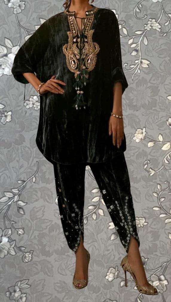 A person wearing a black velvet dress with gold embroidery and tassels, paired with high heels, stands against a gray floral background.
