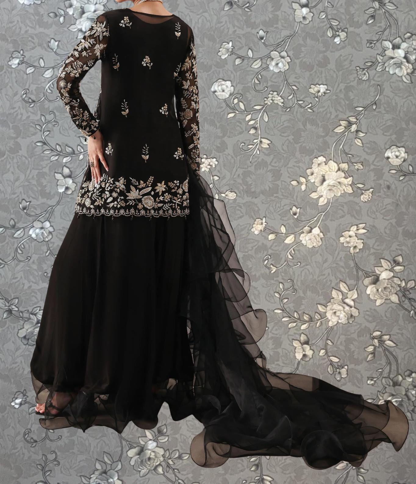A woman wearing a long black dress with intricate embroidery and a flowing train, standing against a gray floral background.