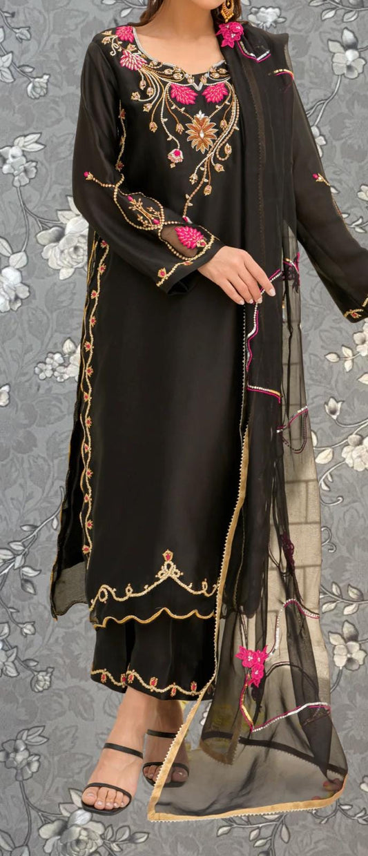 A woman wearing a black dress with intricate embroidery and pink accents, standing against a gray floral background.