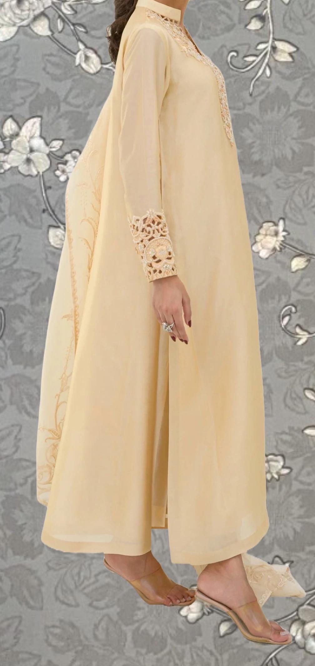 A woman wearing a long, cream-colored dress with lace detailing on the sleeves and neckline, standing against a gray floral background.
