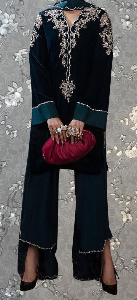 A person wearing a long black dress with intricate embroidery and holding a red clutch purse stands against a gray floral background.