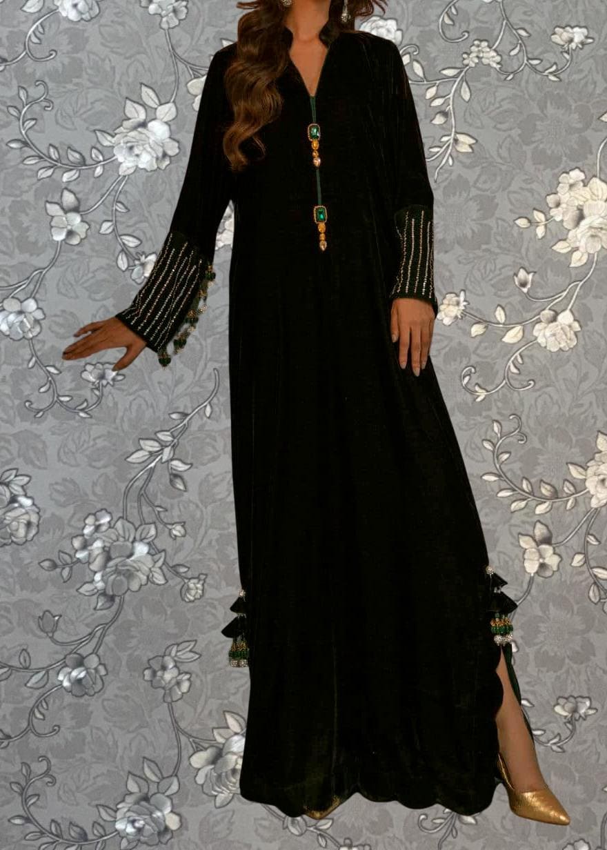 A woman wearing a long black dress with gold accents and tassels, standing against a gray floral background.