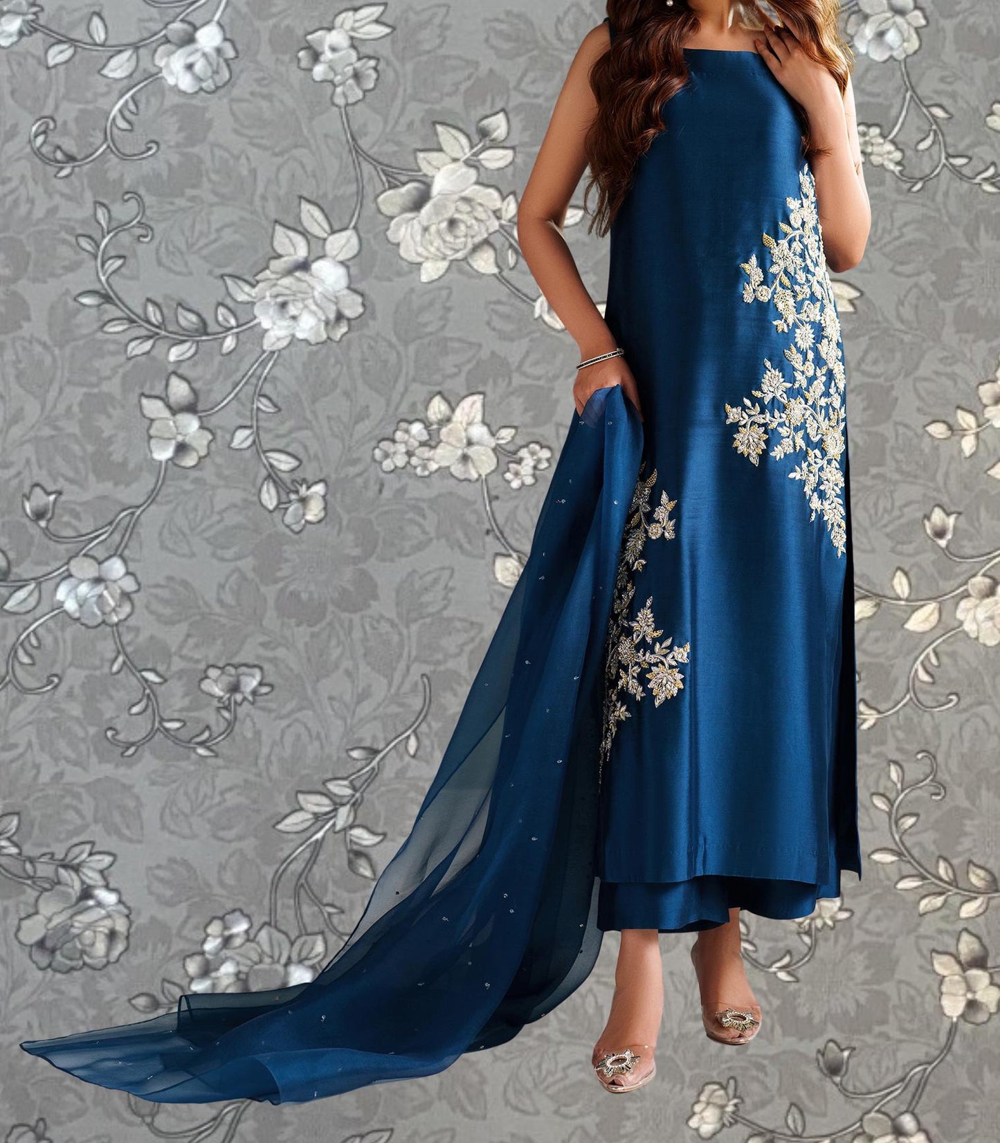 A woman wearing a long blue dress with white floral embroidery stands against a gray floral background.