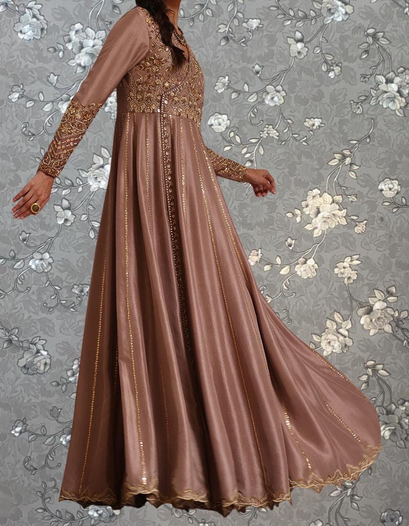 A woman wearing a long, flowing pink dress with intricate gold embroidery stands against a gray floral background.