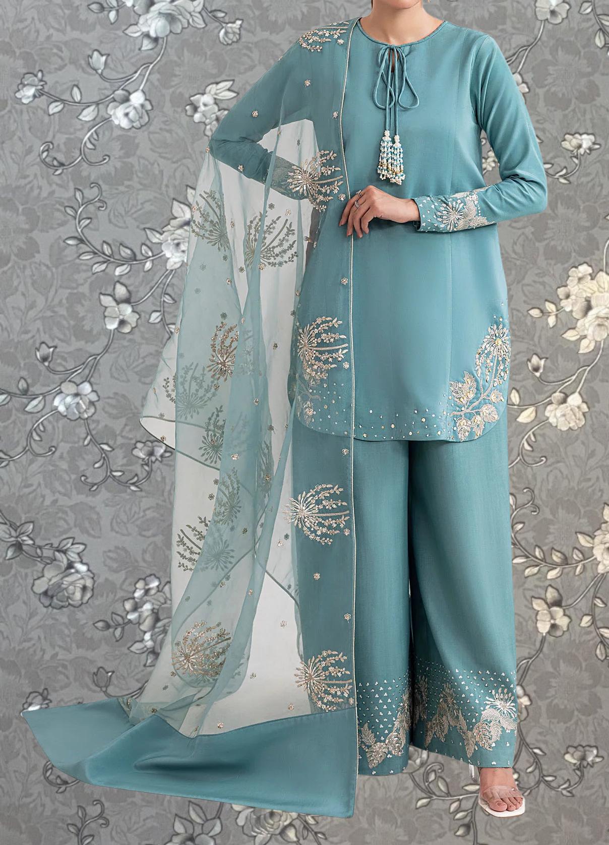 A woman wearing a turquoise dress with intricate embroidered designs, standing against a gray floral background.