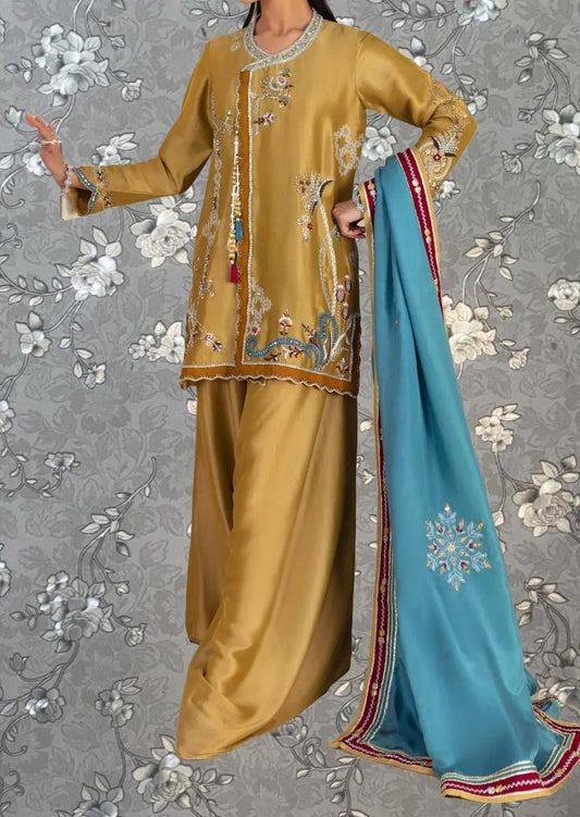 A woman wearing a gold and blue dress with intricate embroidery, standing against a gray floral background.
