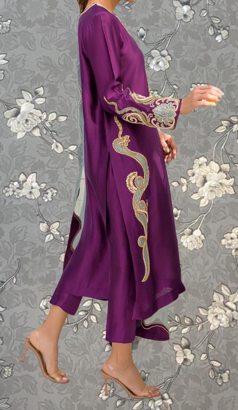 A woman wearing a long purple dress with intricate embroidery stands against a gray floral background.