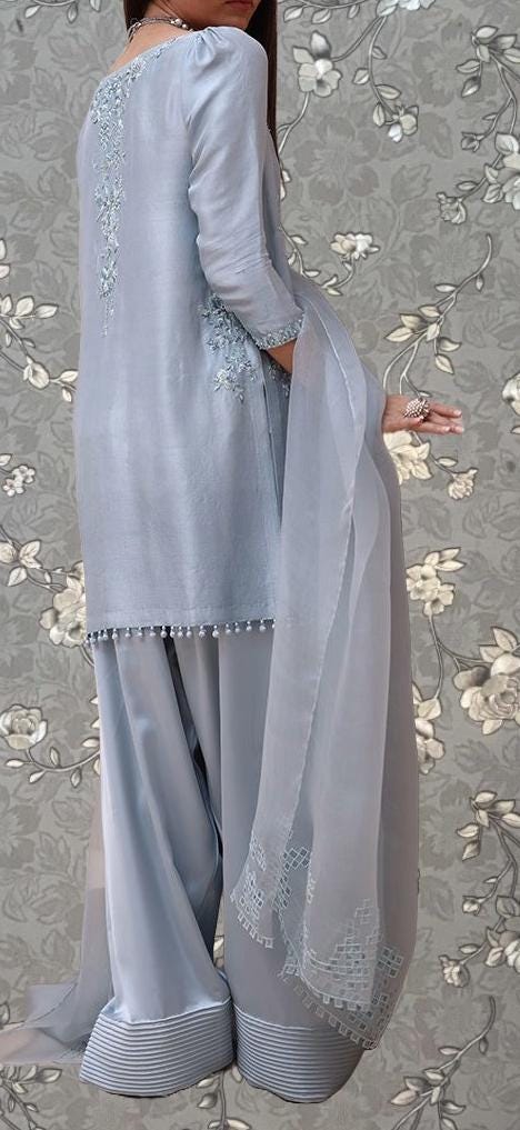 A woman wearing a gray dress with intricate embroidery and a matching gray shawl, standing against a floral patterned background.