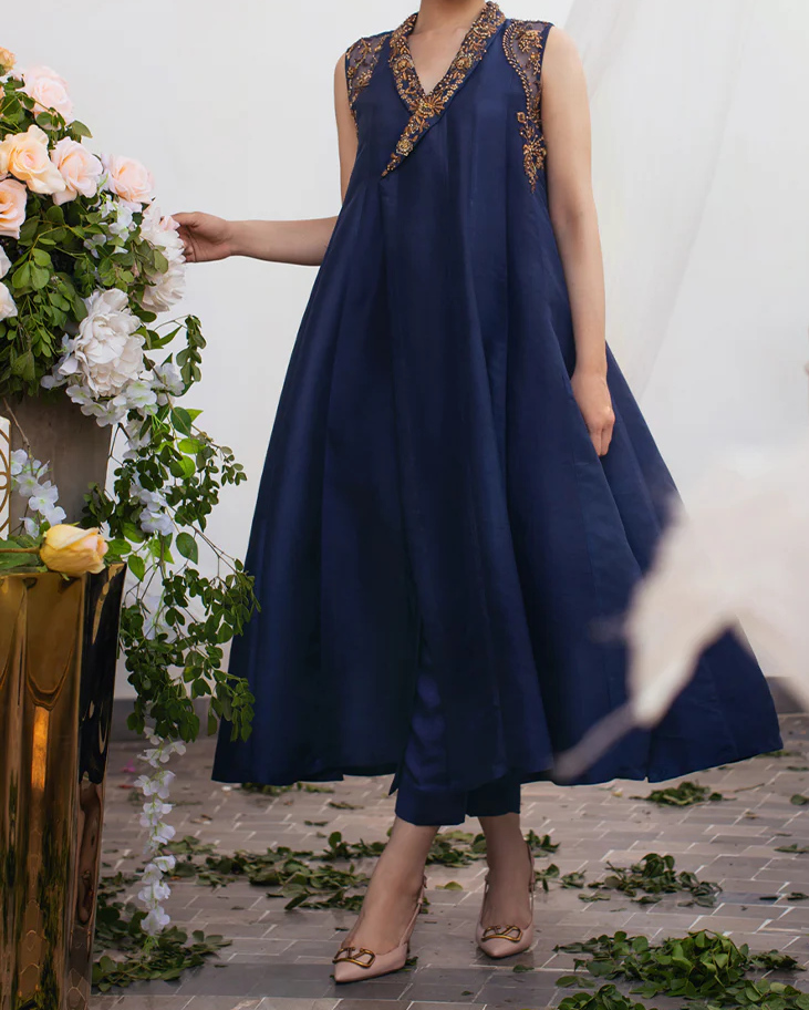 A woman wearing a blue dress with gold accents stands in front of a floral arrangement.