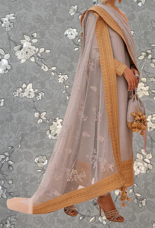 A woman wearing a long, light-colored dress with a gold-colored border and a sheer, embroidered shawl. She is holding a small purse and standing against a gray floral background.