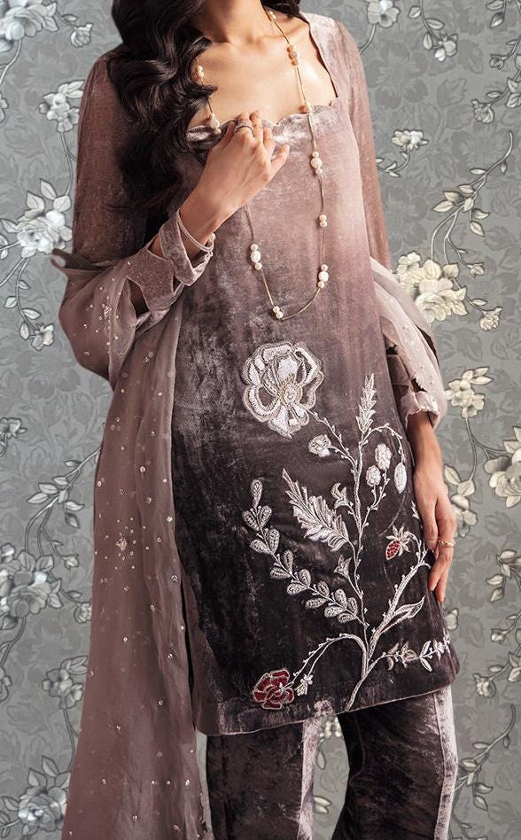 A woman wearing a brown dress with a floral pattern and a long necklace stands against a gray floral background.