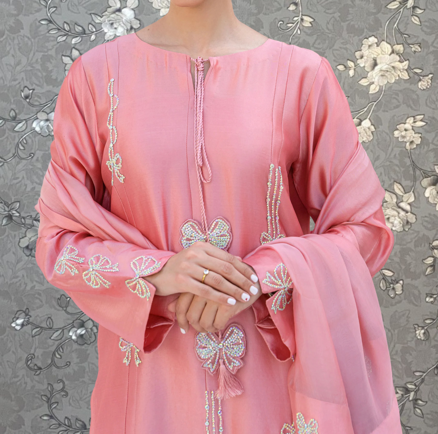 A woman wearing a pink dress with intricate embroidery and floral designs, standing against a gray floral background.