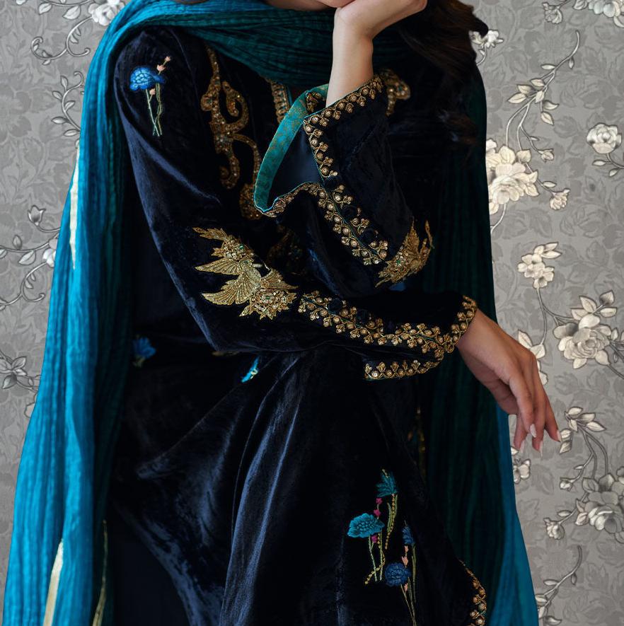 A woman wearing a black velvet dress with gold embroidery and a blue shawl stands against a gray floral background.