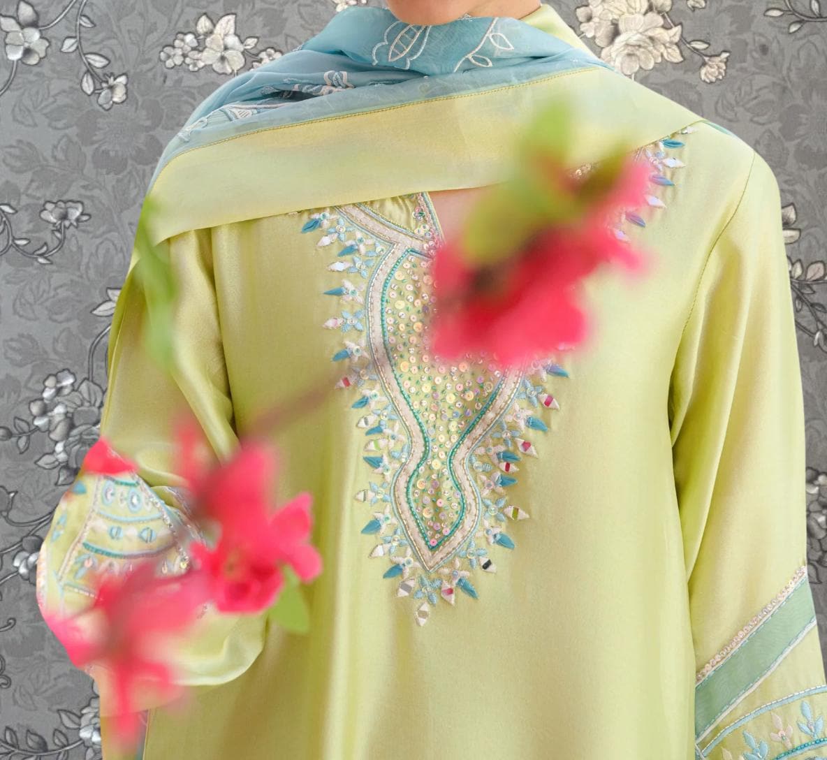 A close-up of a person wearing a yellow dress with intricate embroidery and a blue scarf, with blurred pink flowers in the foreground.