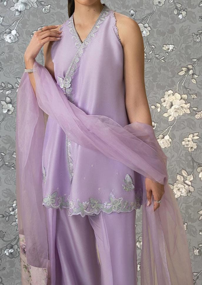 A woman wearing a lavender dress with a matching purple shawl stands against a gray floral background.