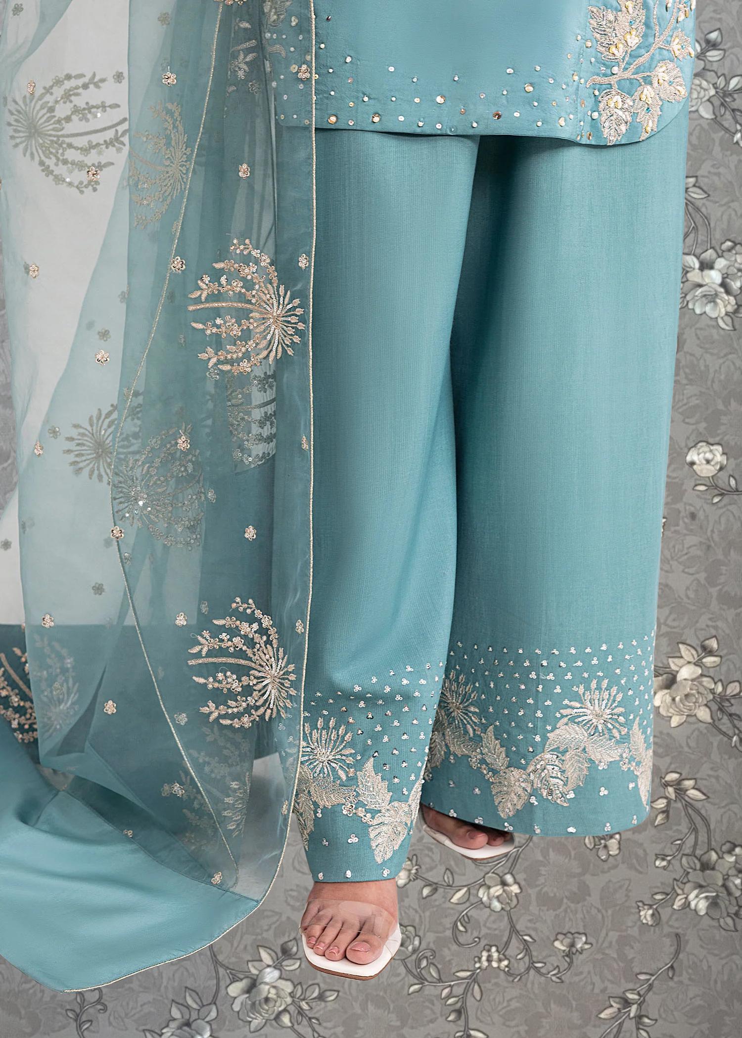 a person wearing a pair of blue pants with intricate embroidered designs on the fabric. The pants are paired with a matching blue and white patterned shawl. The background features a floral pattern, which complements the overall design of the outfit.