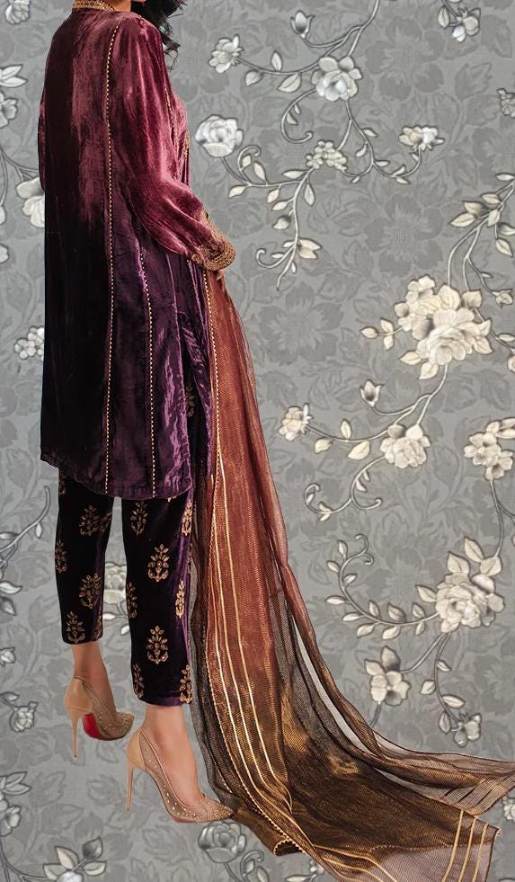 A person wearing a long purple velvet dress with gold floral patterns, standing against a gray floral background.