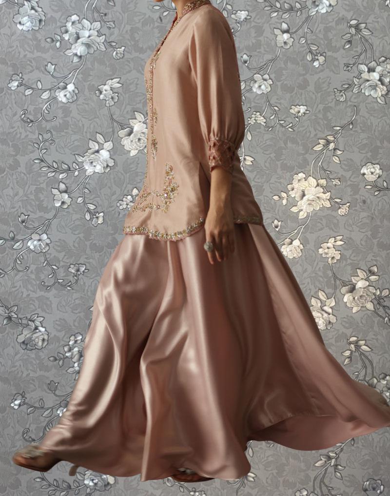 A woman wearing a long, flowing pink dress with intricate embroidery stands against a gray floral background.