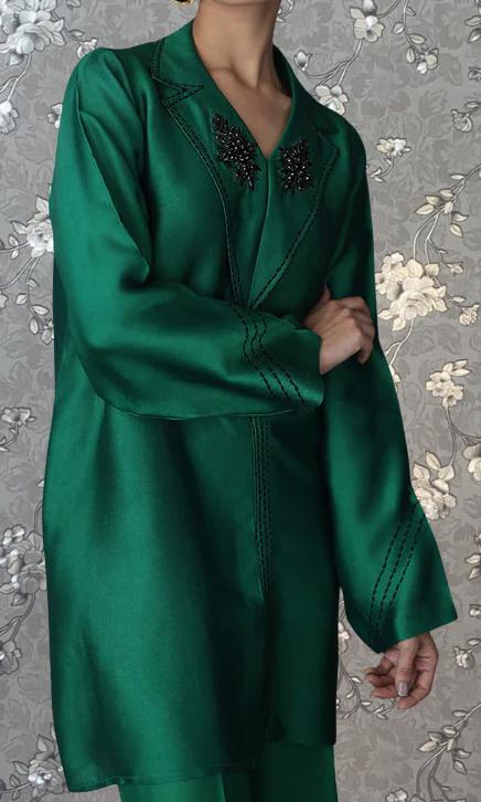 A person wearing a long green coat with black embellishments on the collar and sleeves stands against a floral background.