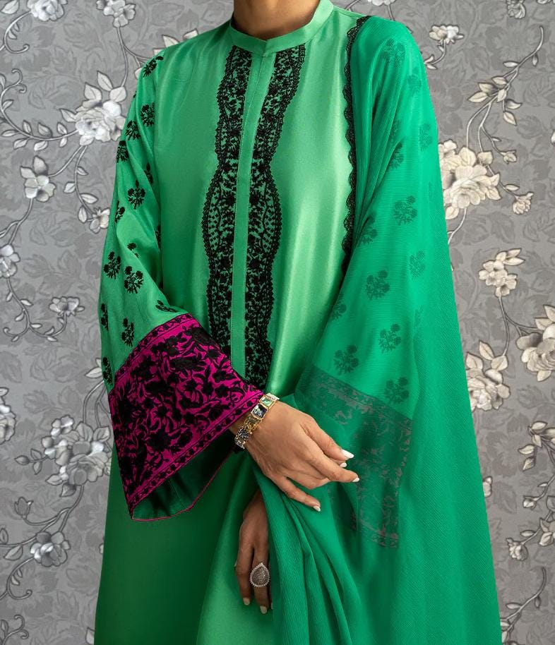 A person wearing a green dress with pink accents and black embroidery stands against a gray floral background.