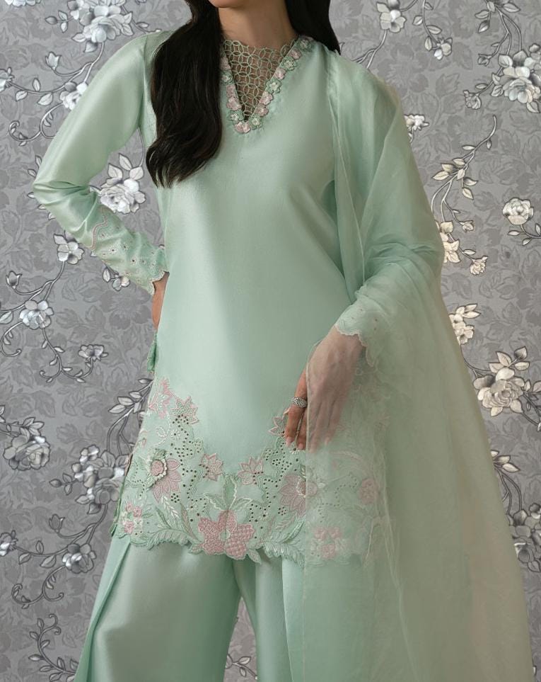 A woman wearing a mint green dress with intricate embroidery and floral patterns, standing against a gray floral background.