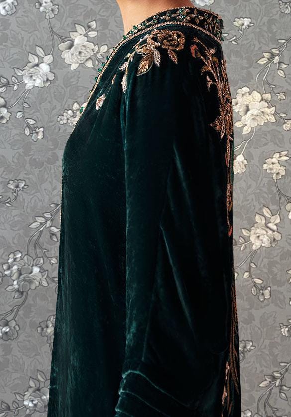 A close-up of a dark green velvet dress with gold embroidery on the sleeves and collar, standing against a gray floral background.