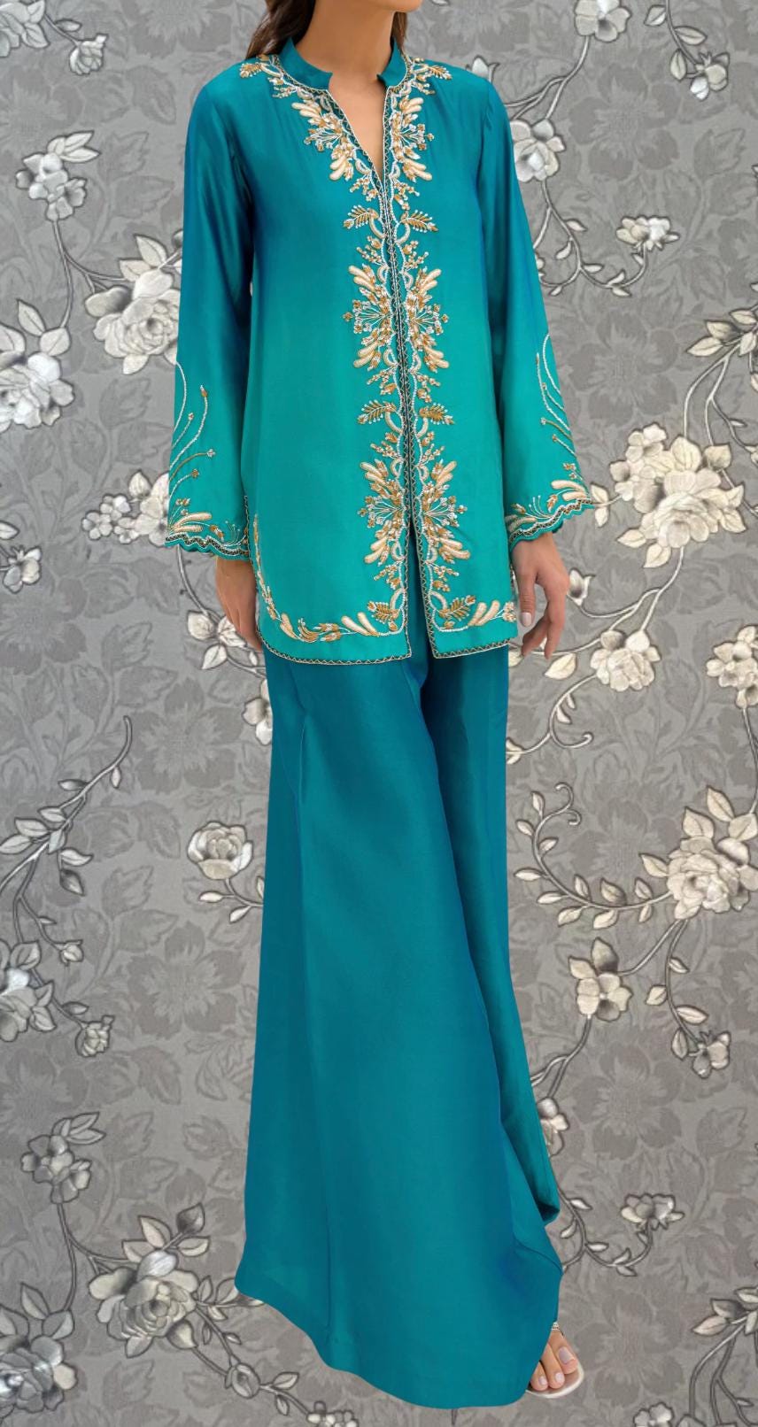 A woman wearing a turquoise dress with gold embroidery stands against a gray floral background.